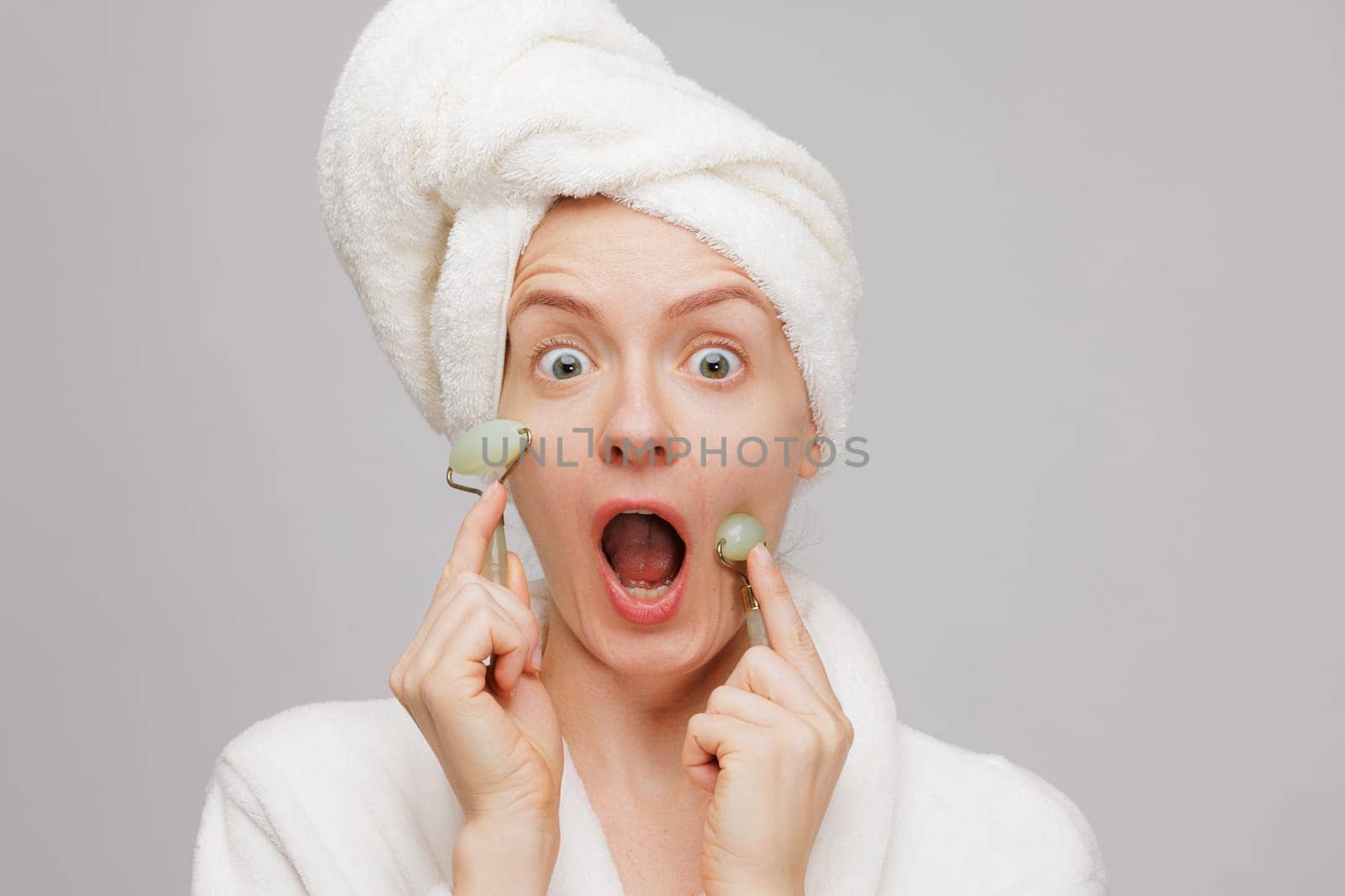 A surprised woman with a towel on her head and a bathrobe, holding jade rollers on her cheeks. She has wide eyes and an open mouth, expressing excitement or shock.