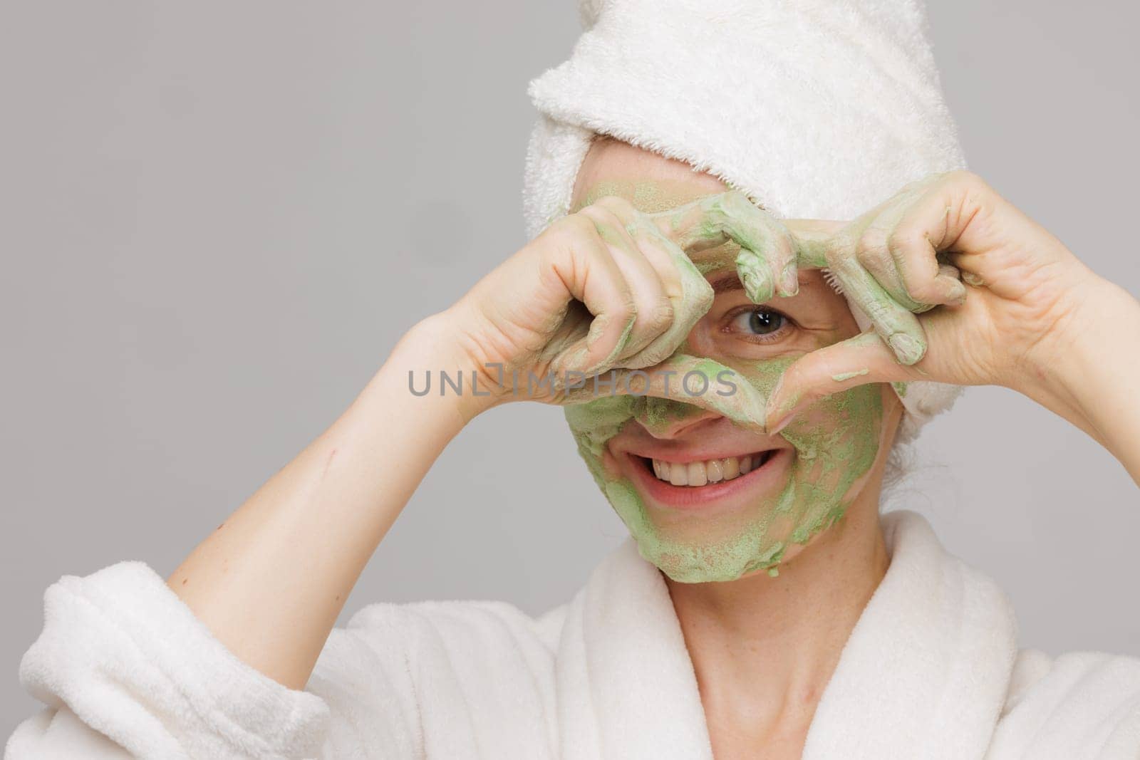 A woman with a towel on her head smiles as she applies a green face mask. She has a happy expression on her face, showing off her skincare routine