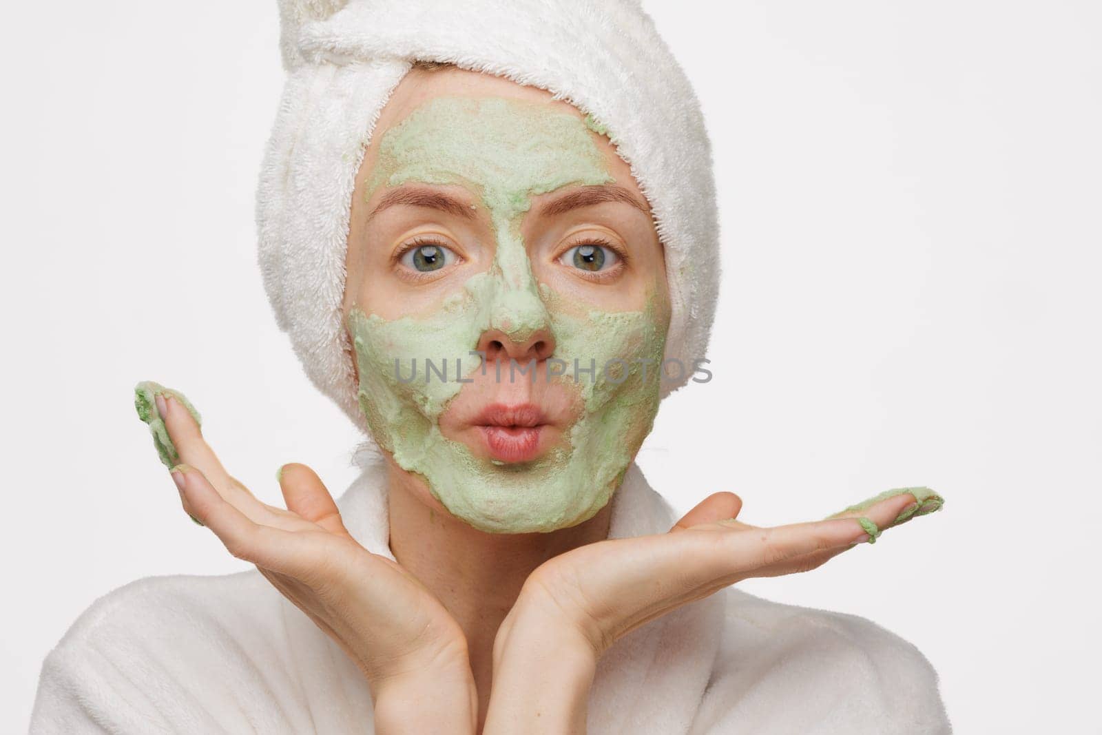 A woman with a towel on her head smiles as she applies a green face mask. She has a happy expression on her face, showing off her skincare routine