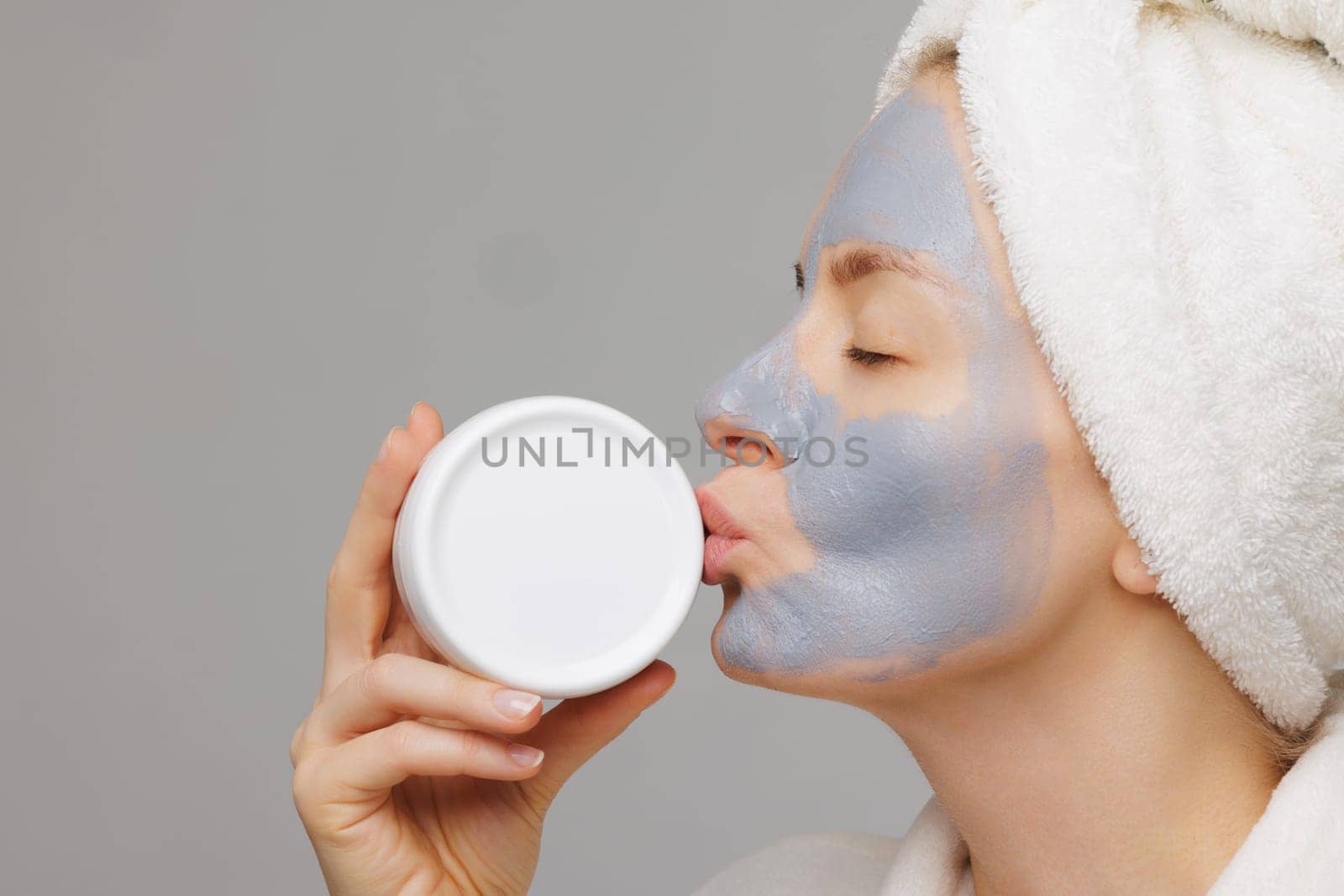Woman with mask on her face playfully kisses her favorite white jar of cream. concept of skin care for face and body