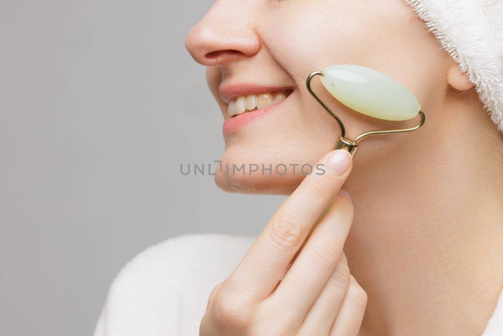 A woman in a white robe and a towel on her head smiles while holding jade facial rollers under her eyes.. wrinkle smoothing concept