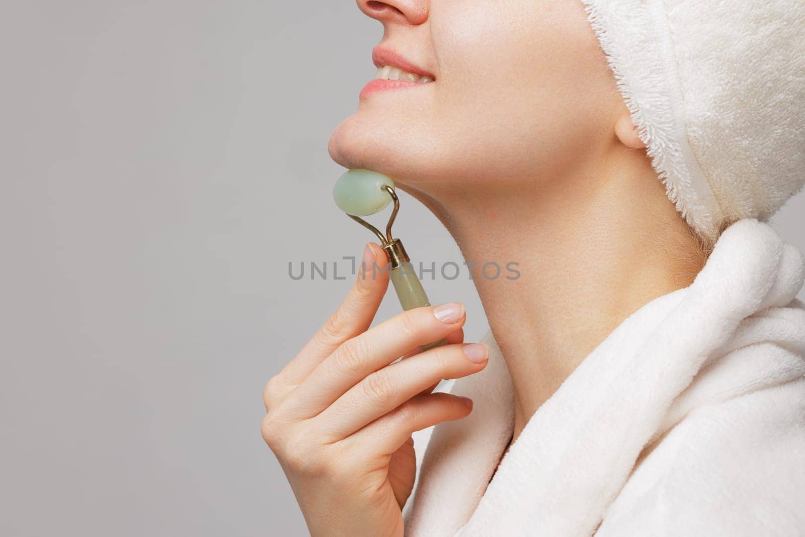 Woman using jade roller on her chin, wearing white robe and towel on her head. skin care concept. facial skin smoothing and massage