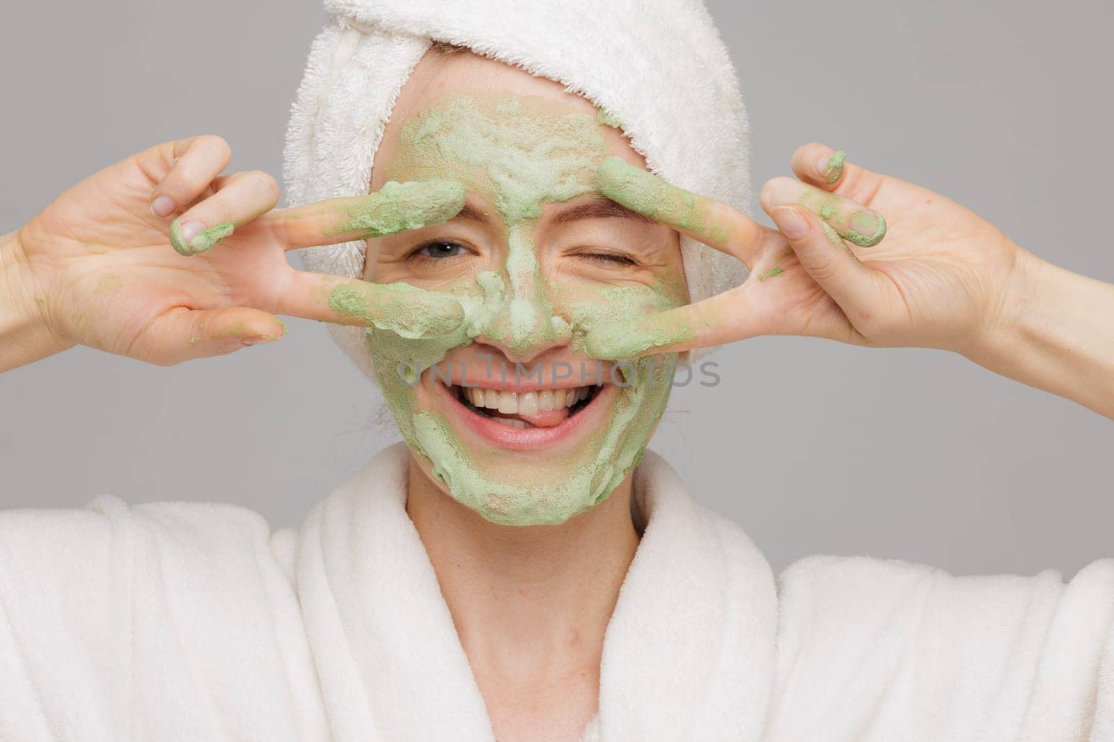 A woman with a towel on her head smiles as she applies a green face mask. She has a happy expression on her face, showing off her skincare routine