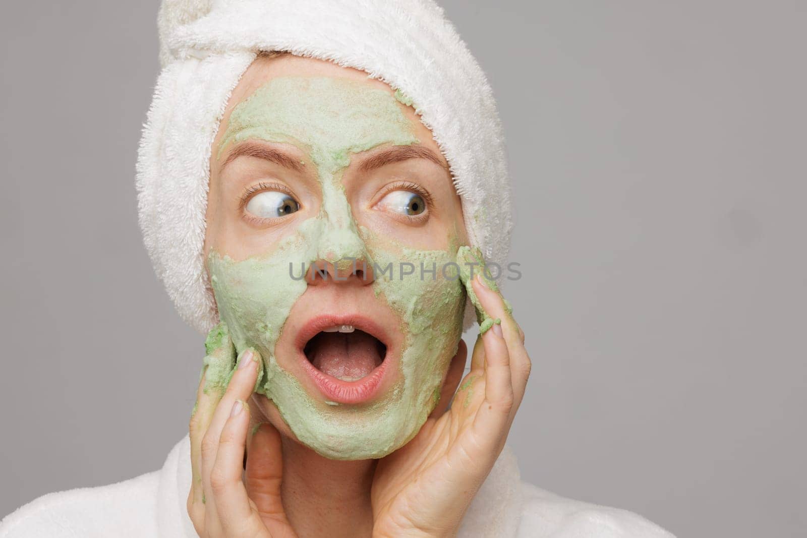 A woman with a towel on her head smiles as she applies a green face mask. She has a happy expression on her face, showing off her skincare routine