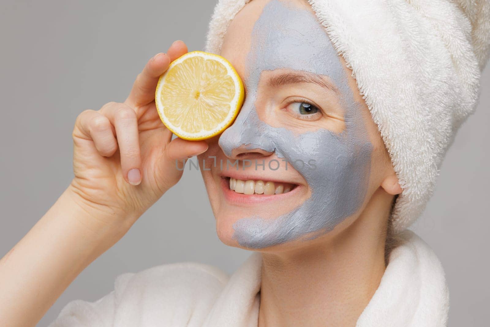 A woman with a mask on her face holding a lemon wedge in front of her mouth. She is wearing a white robe and a towel on her head. Beauty and Skin care concept
