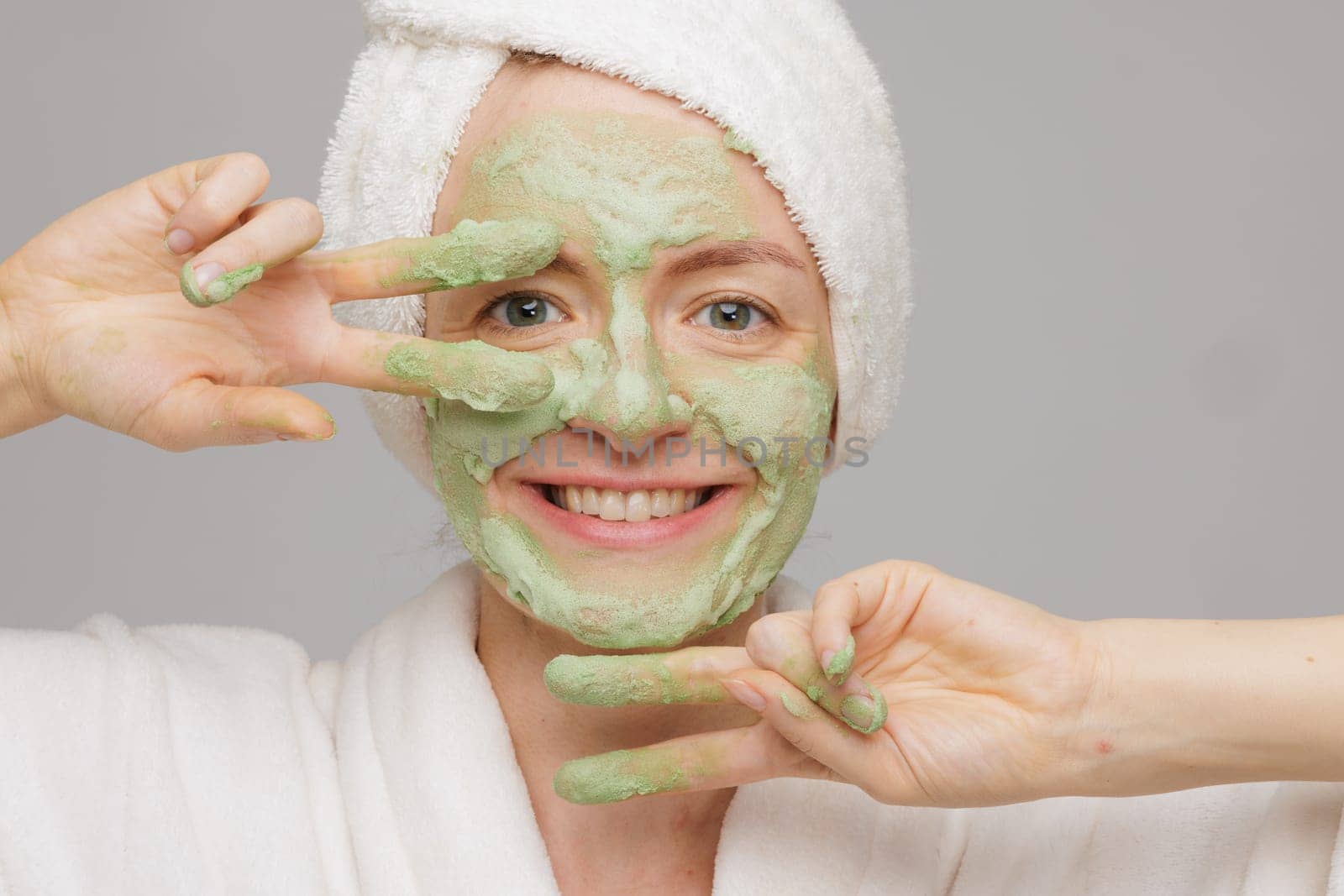 A woman with a towel on her head smiles as she applies a green face mask. She has a happy expression on her face, showing off her skincare routine
