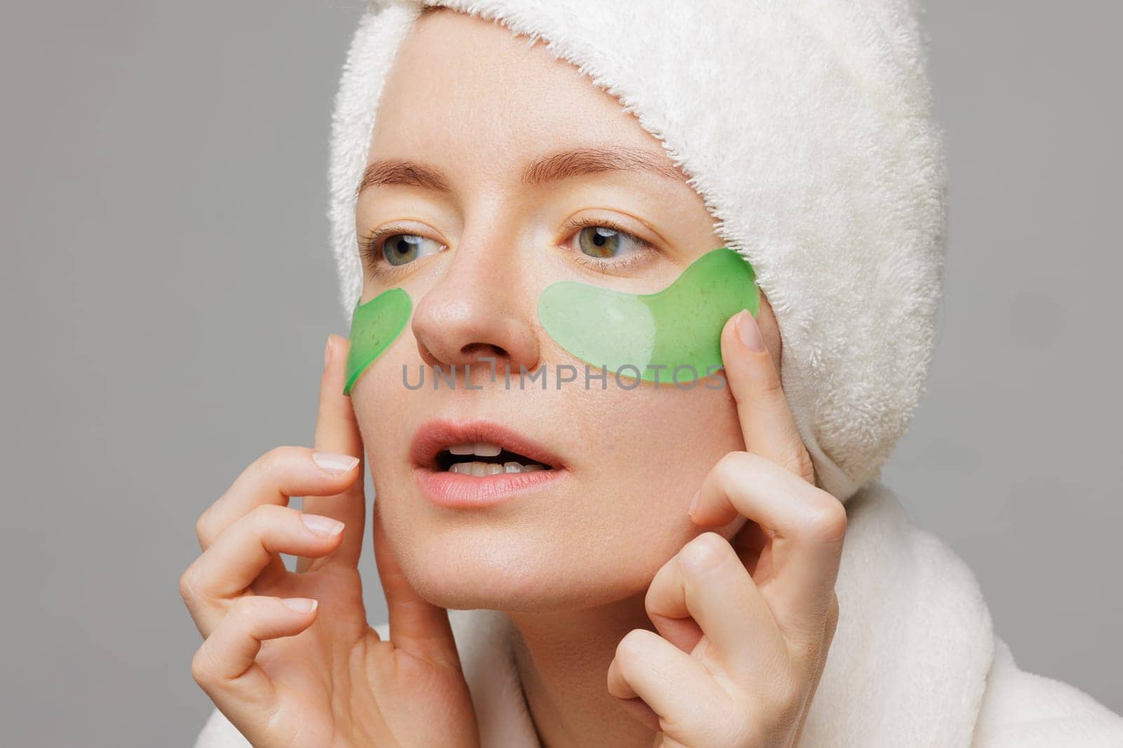 A woman with a towel on her head and a robe smiles as she applies green gel patches under her eyes facial cosmetology concept