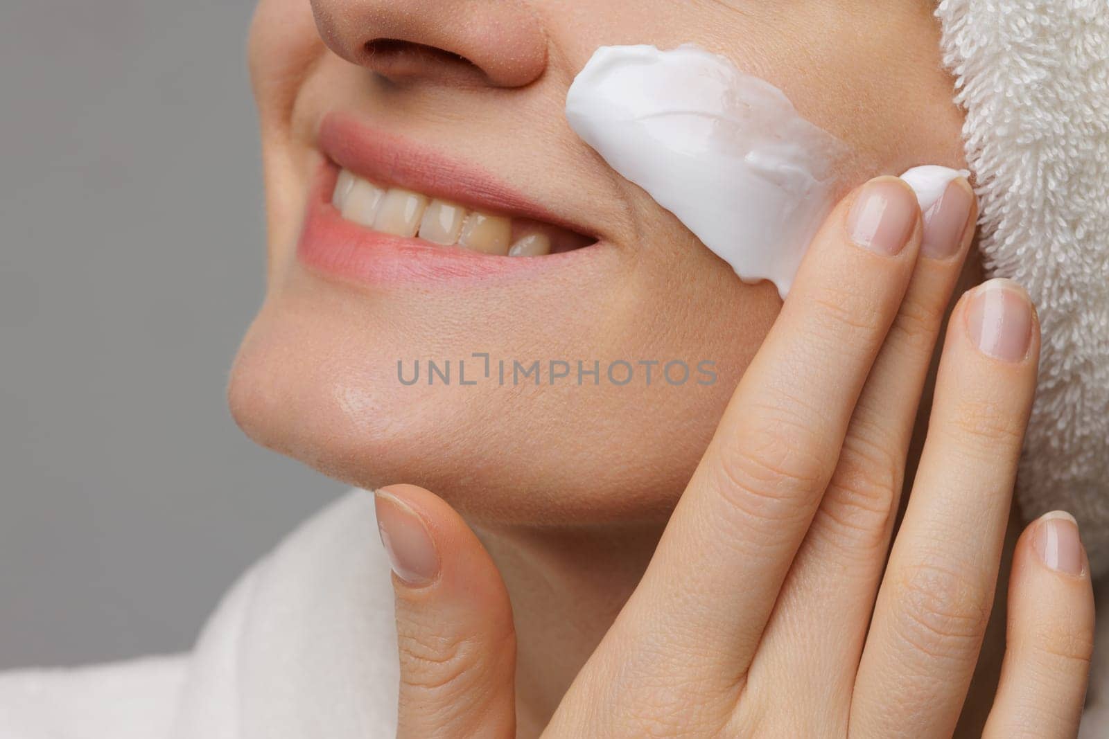 A woman applying cream to her face, smiling gently. She has a towel wrapped around her head and is in a relaxed setting, suggesting a skincare routine or spa treatment.