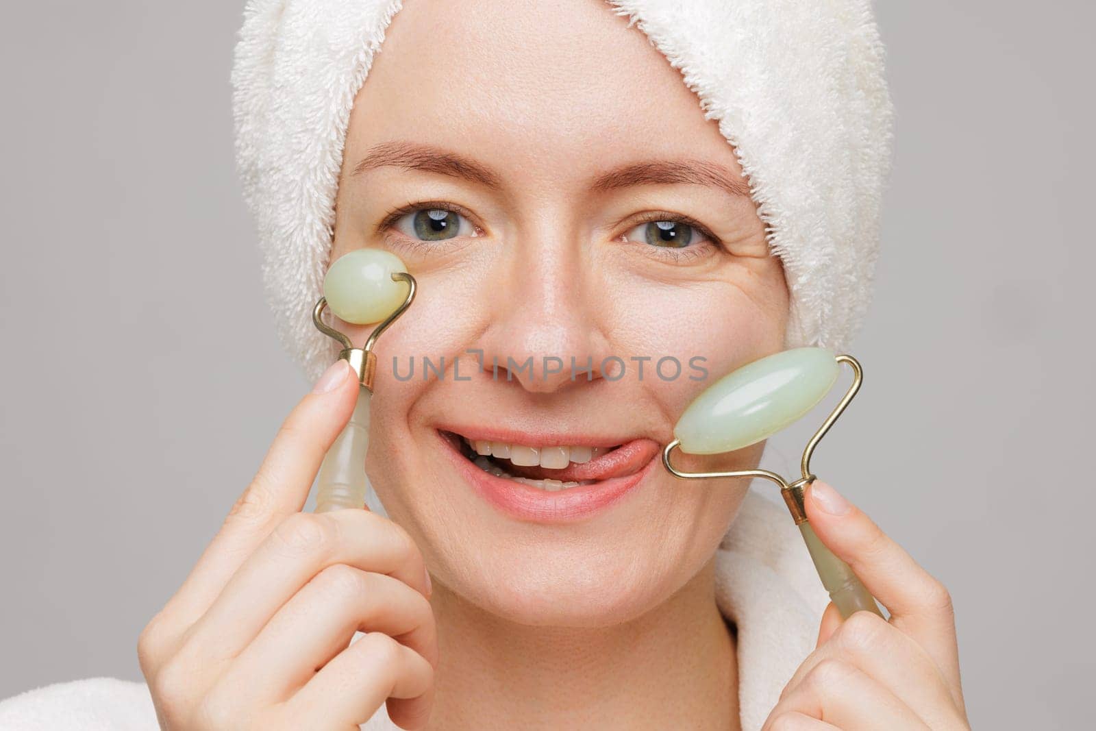 A woman with a towel on her head and a robe playfully uses jade facial rollers on her cheeks. She has a surprised expression, concept skin care facial.