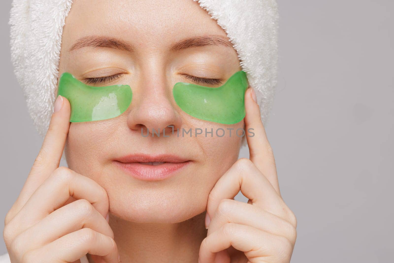 A woman with a towel on her head and a robe smiles as she applies green gel patches under her eyes facial cosmetology concept