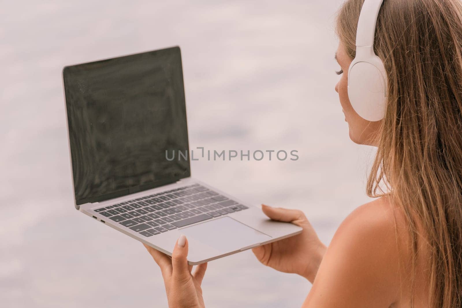 A woman is sitting on a beach with a laptop in her lap. She is wearing headphones and she is listening to music
