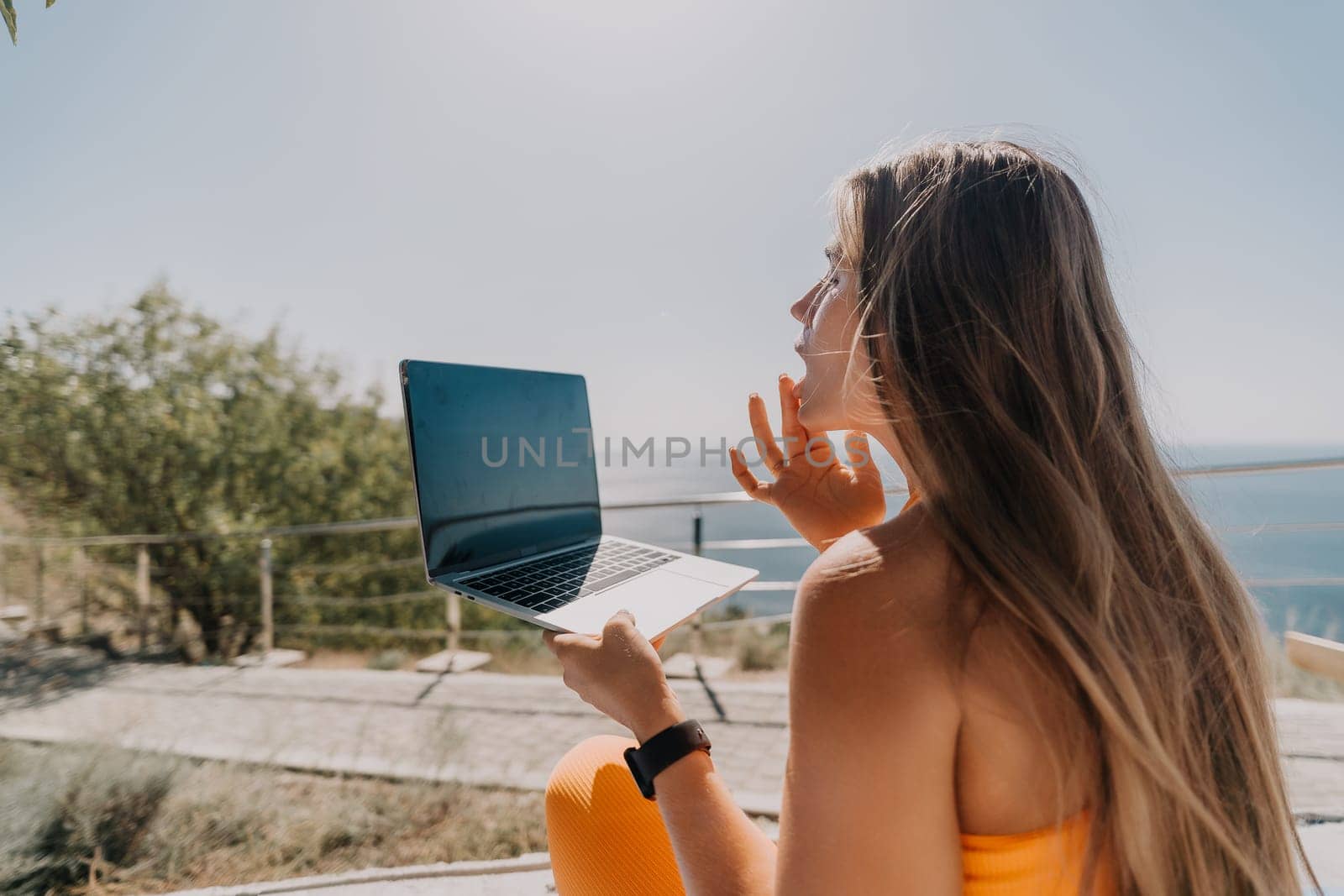 A woman with long blonde hair sits outdoors and uses a laptop. She is looking at the screen and has a thoughtful expression.