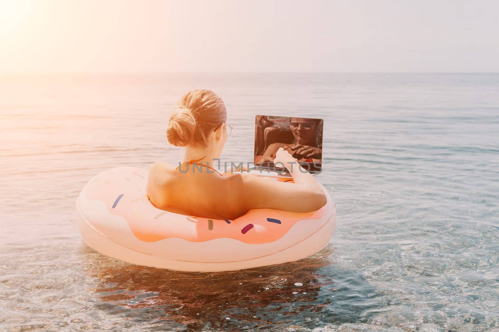 Woman freelancer works on laptop swimming in sea on pink inflatable ring. Pretty lady typing on computer while floating in the sea on inflatable donut at sunset. Freelance, remote work on vacation