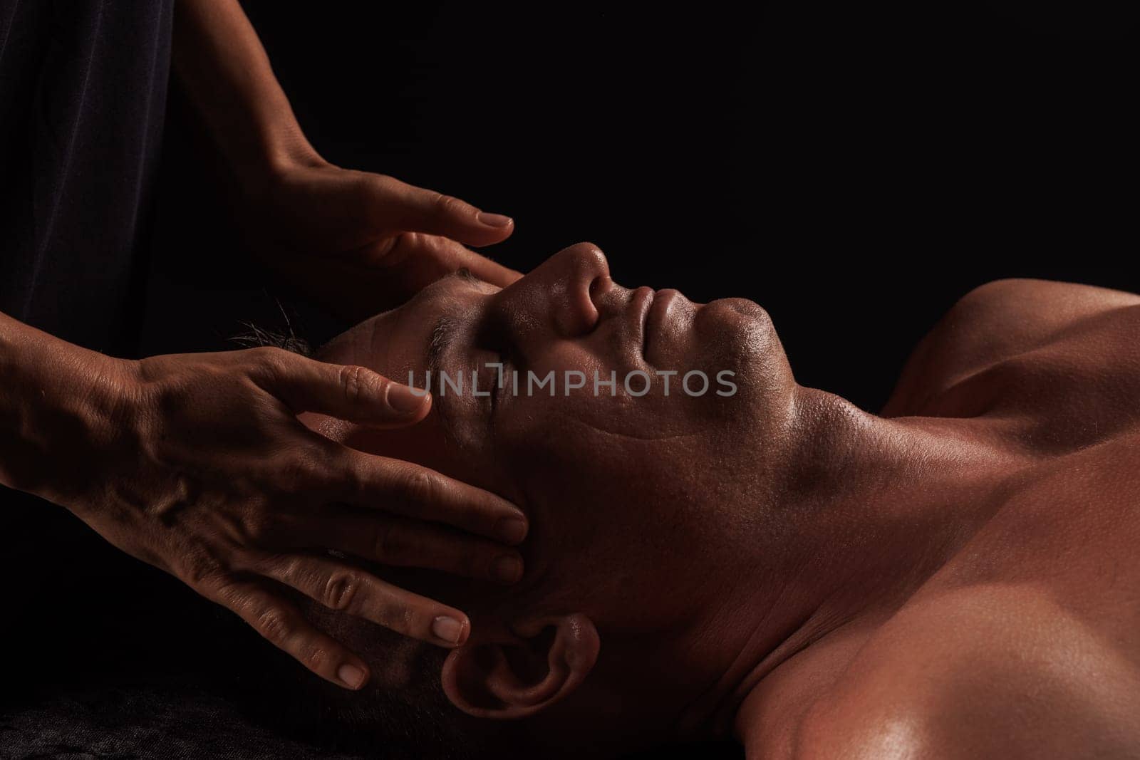 Close-up of a man receiving a facial massage against a dark background, The hands of the massage therapist are gently pressing on his forehead and temples.