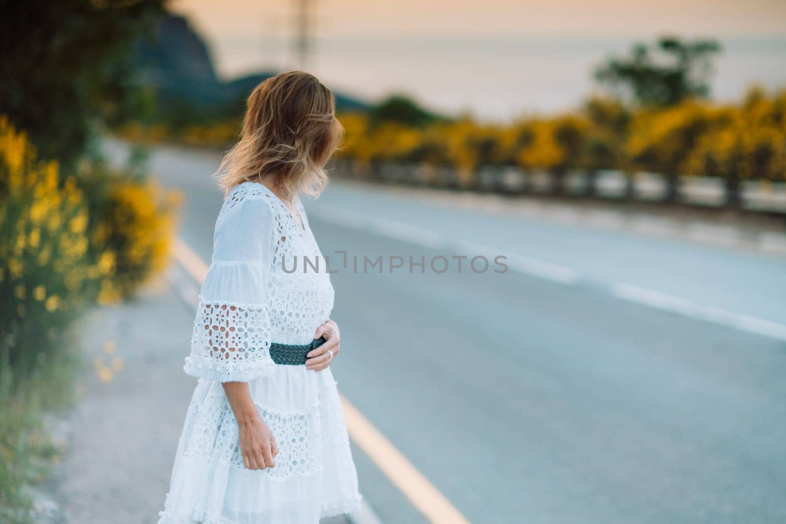 A woman in a white dress is standing on the side of a road. She is looking off into the distance, possibly lost in thought. The scene is peaceful and serene, with the woman's dress