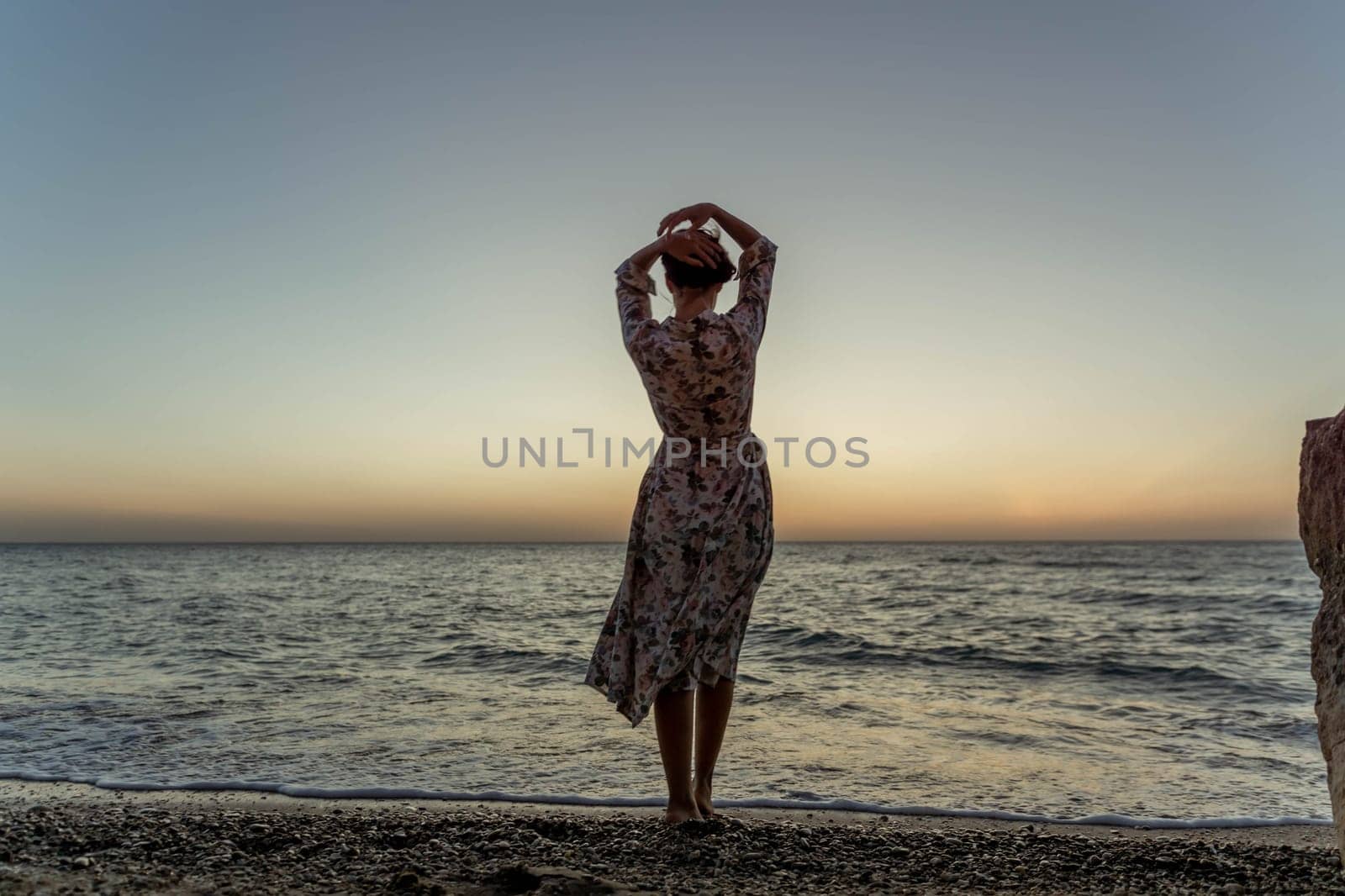 A woman in a dress stands on a beach, facing the ocean, with her arms raised above her head as the sun sets.