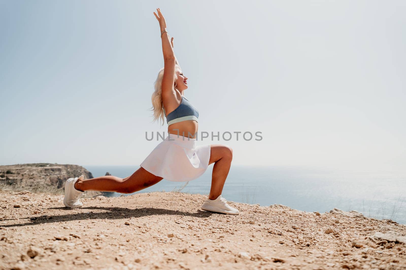 A blonde woman in a white skirt and blue sports bra performs a stretching exercise on a cliff overlooking the sea.
