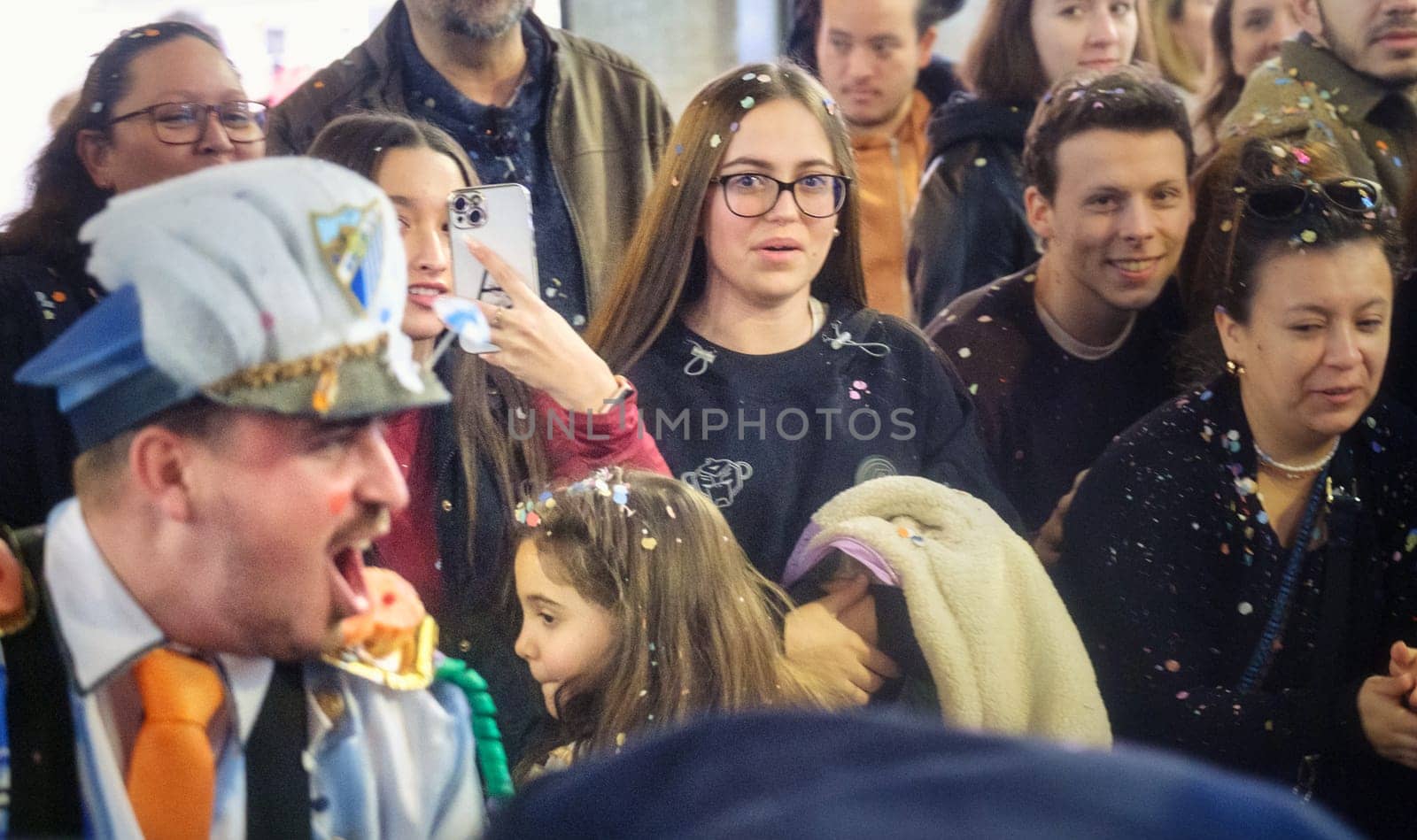 In this image, you see a group of people gathered together in what appears to be a public event or celebration. In the foreground, a person is dressed in costume, which includes a hat that looks like it might be part of a uniform, and they are making a dramatic expression with their mouth open wide...To the right of the image, there's a young woman with glasses looking toward the camera. She's wearing a black top with some pattern or design on it, and there are specks of what could be confetti in her hair and on her clothes, suggesting the festive nature of the event...Other individuals in the scene are also adorned with confetti, and some are holding items that might be part of the event or celebration. The mood overall is joyful and lively, and it seems like they are all watching or participating in some kind of performance or activity.