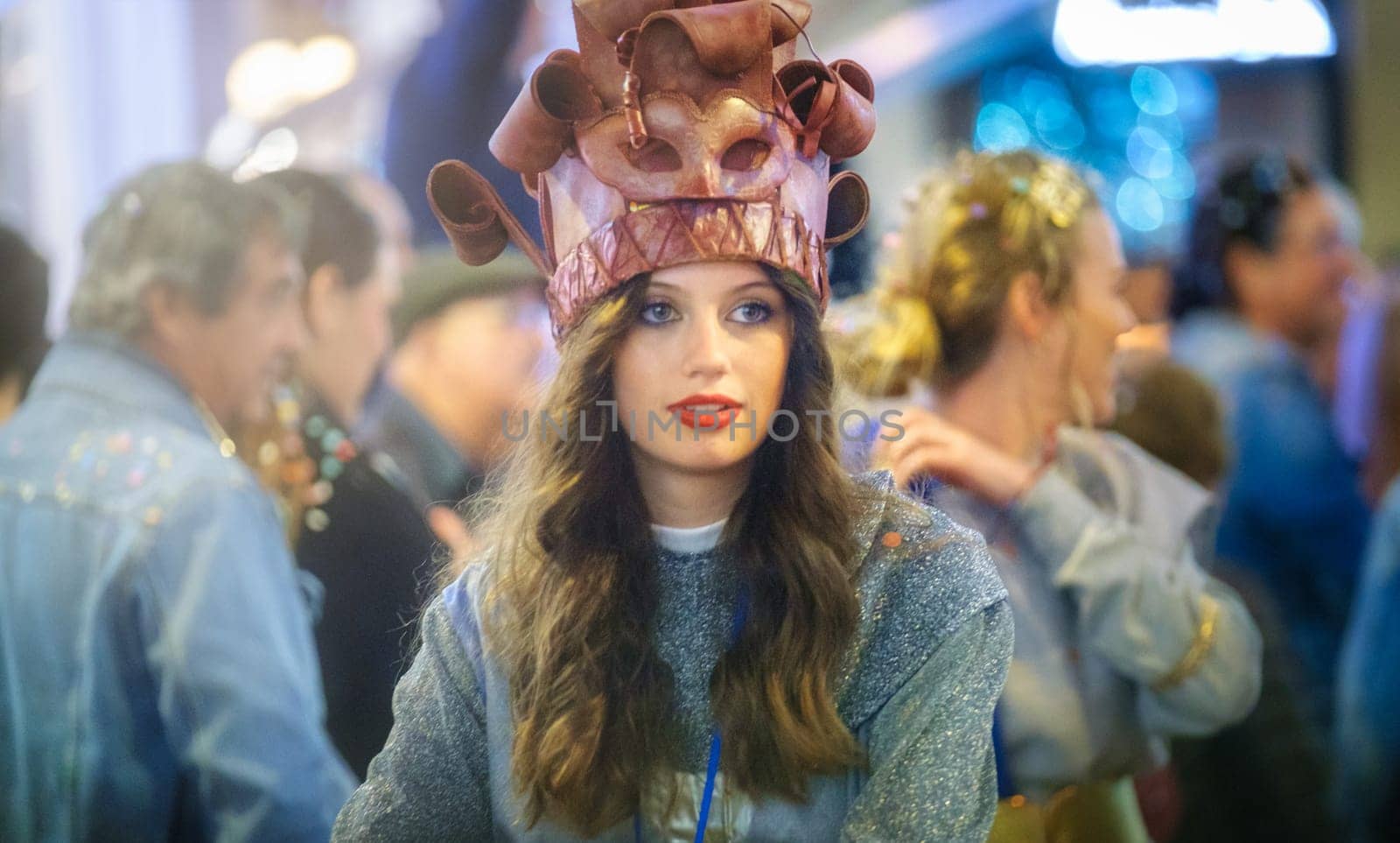 In the image, there is a person wearing an unusual hat that appears to be crafted to resemble a cake or dessert with rolling pins as decorations. The person has long hair, red lipstick, and is looking directly at the camera with a neutral expression. They are wearing a sweater or top with a shimmering texture, and behind them, there are blurred figures of other people, suggesting that the photo was taken at an event or a gathering with a fair amount of people. The lighting suggests an indoor setting with artificial illumination.
