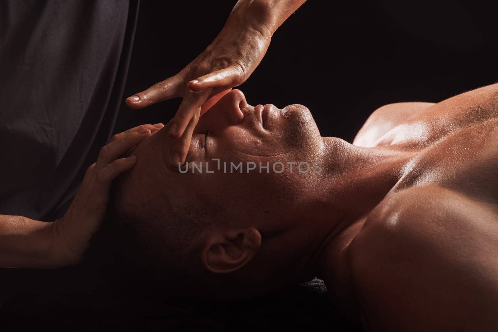 Close-up of a man receiving a facial massage against a dark background, The hands of the massage therapist are gently pressing on his forehead and temples.