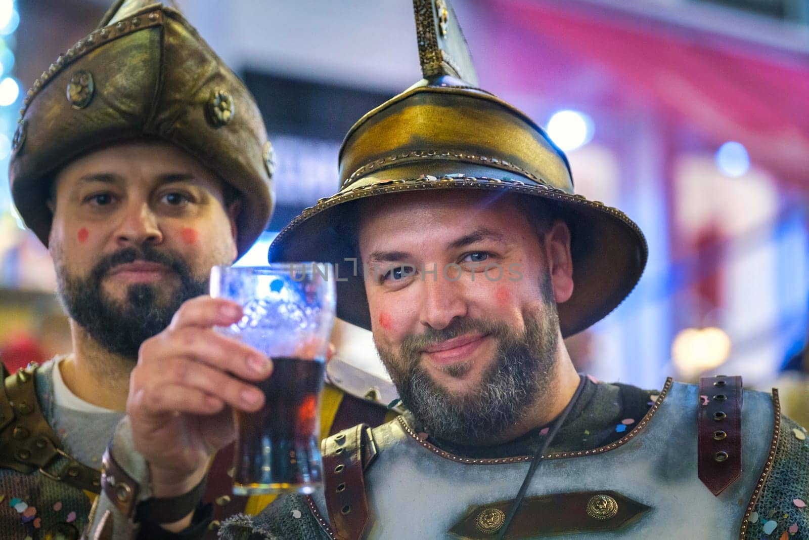 In the image, there are two individuals who appear to be dressed in costume, reminiscent of historical or fantasy armor. Both are wearing metal helmets with a peak or spike at the top, and the attire includes what looks like metallic and studded shoulder guards, suggesting a knight or warrior type of theme...The person on the right is smiling and holding up a glass with a dark liquid, possibly beer, in a toasting gesture. The other individual is also smiling but isn't directly engaging with the camera. They both have red marks on their cheeks, which could be part of the costume effect or some kind of celebration markings. The setting appears to be a festive or social environment, likely a place where dressing up and enjoying beverages is part of the event's atmosphere.