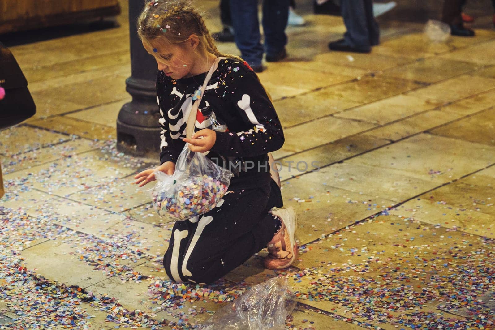 The image shows a young girl kneeling on the floor surrounded by colorful confetti. She appears to be holding onto a clear plastic bag that contains more confetti, suggesting that she may have been collecting it or that the bag has spilled. Her focus and the way she's interacting with the confetti give the impression that she is either playing or cleaning up. The environment looks like an indoor venue, possibly after a celebration or an event where confetti was used. The scattered confetti adds a vibrant touch to the image, reflecting a festive or celebratory atmosphere.