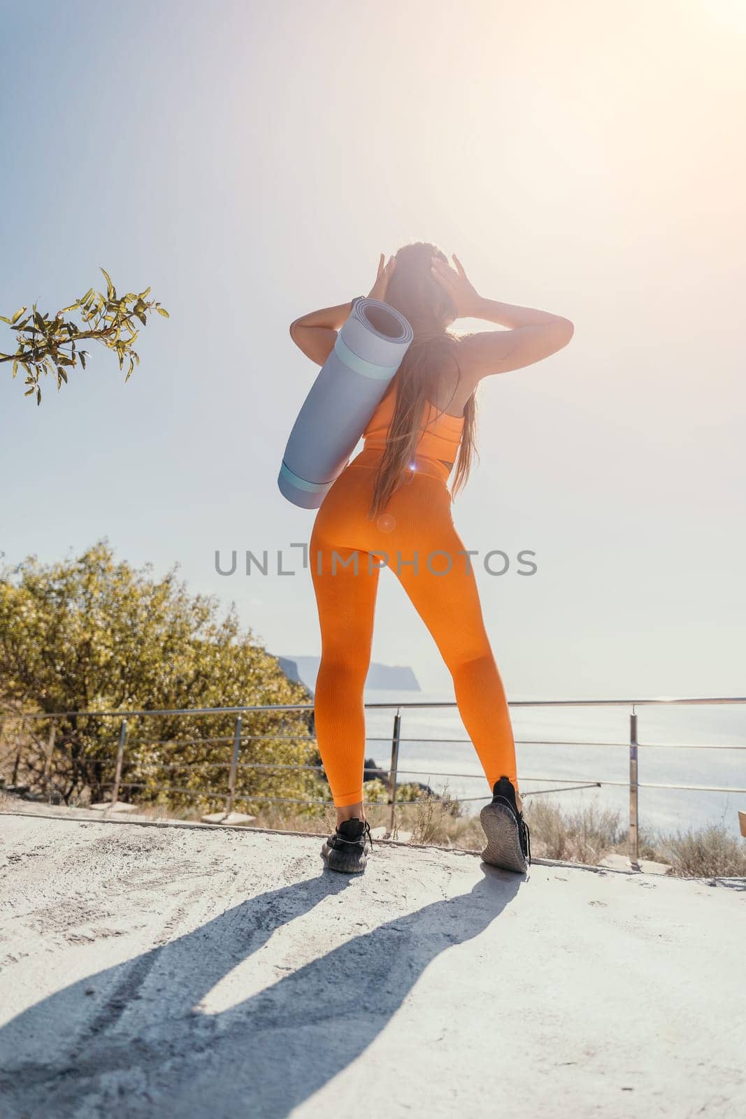 A woman wearing an orange yoga outfit with a blue yoga mat on her back is standing on a concrete surface, looking out at the sea.