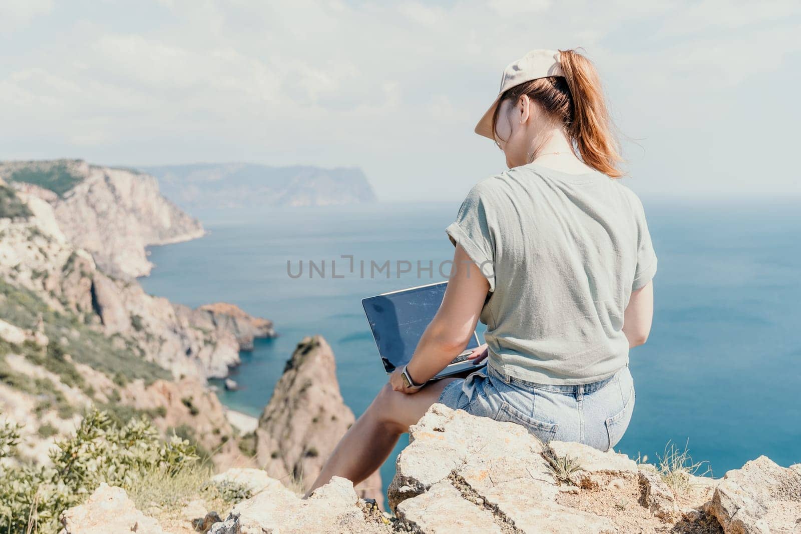Digital nomad, woman in the hat, a business woman with a laptop sits on the rocks by the sea during sunset, makes a business transaction online from a distance. Freelance, remote work on vacation.