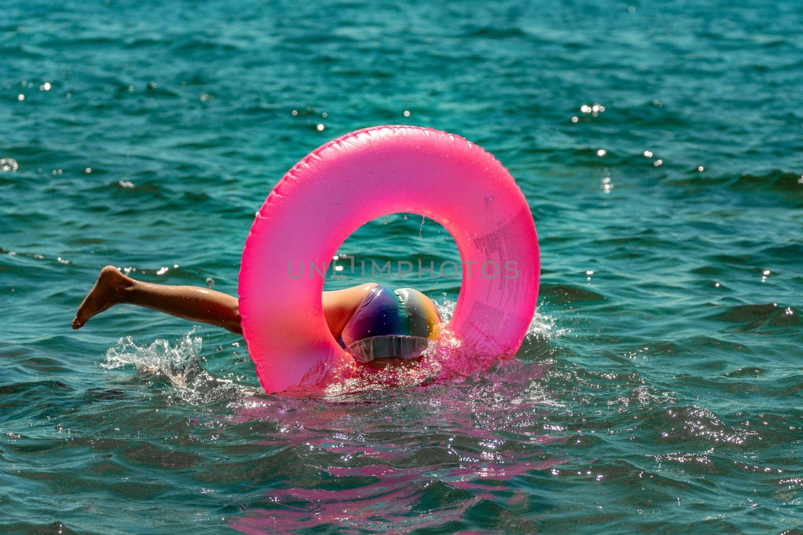 A child wearing a blue and white hat floats on a pink inflatable ring in the blue ocean. They are upside down and submerged.