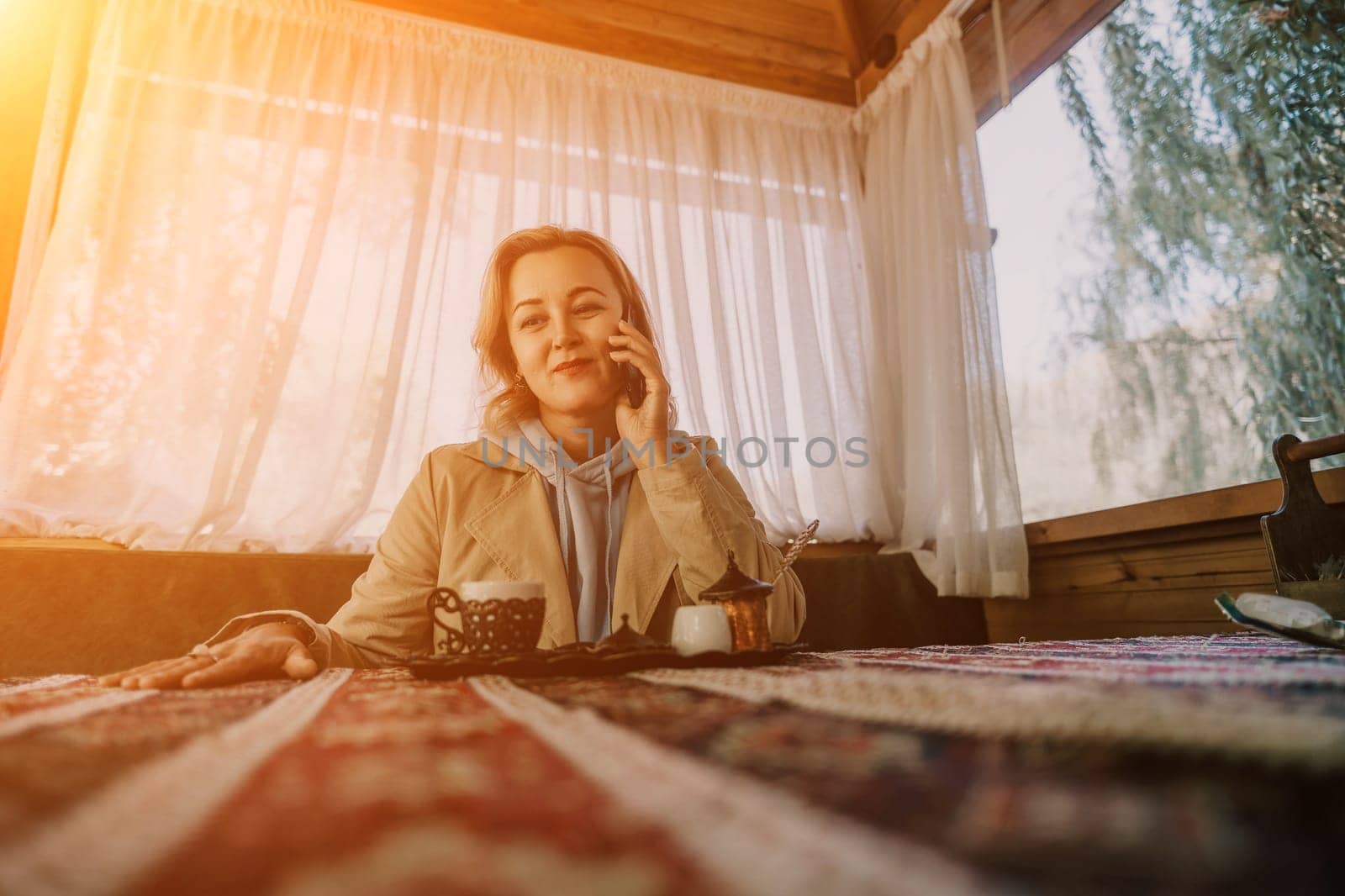 A woman sits at a table in a wooden gazebo, talking on a cell phone. Sunlight streams through the window behind her.