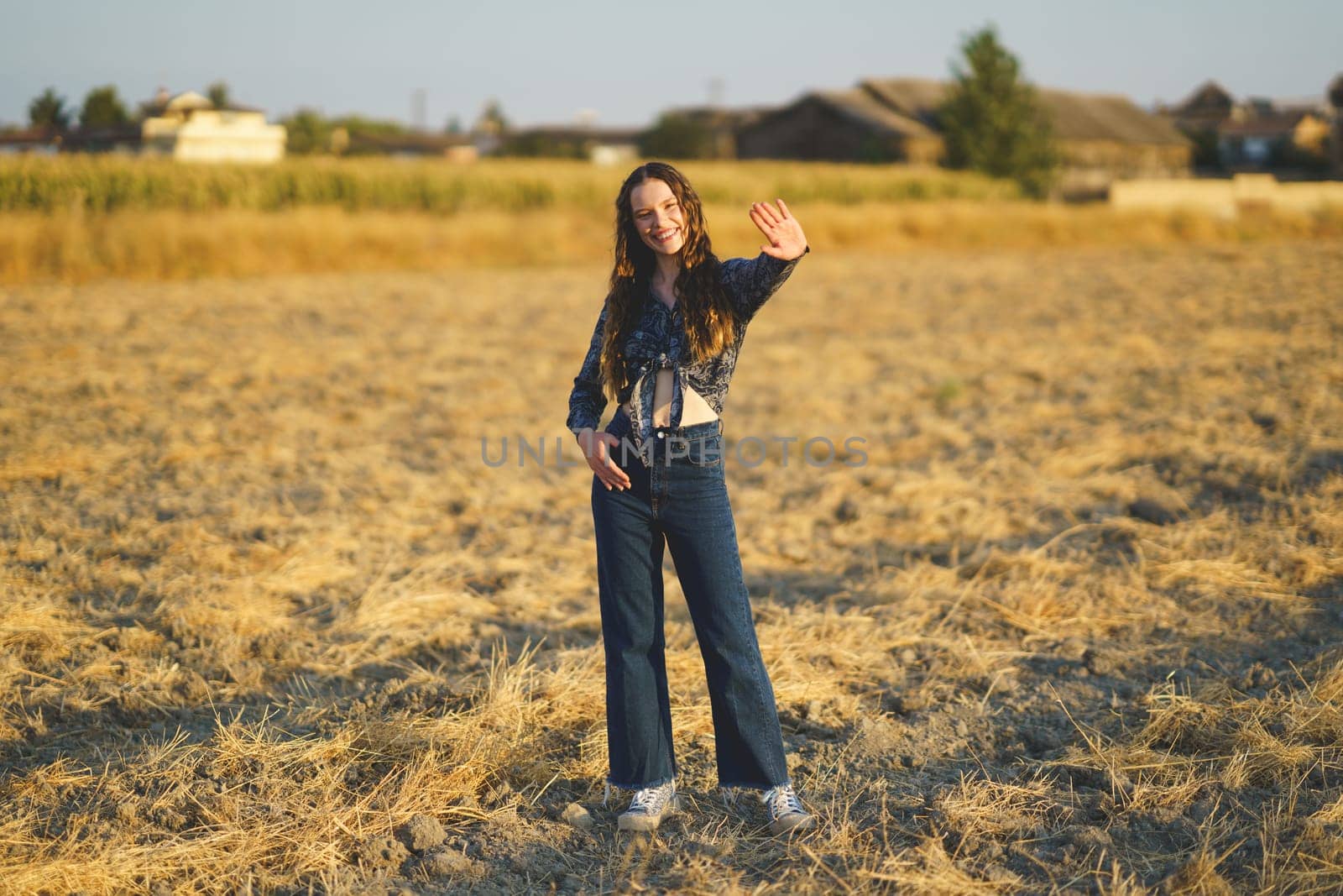 In a sunlit golden field, a young woman beams with joy as she waves cheerfully, dressed in a trendy denim outfit that perfectly embodies a warm and relaxed vibe, radiating happiness and freedom