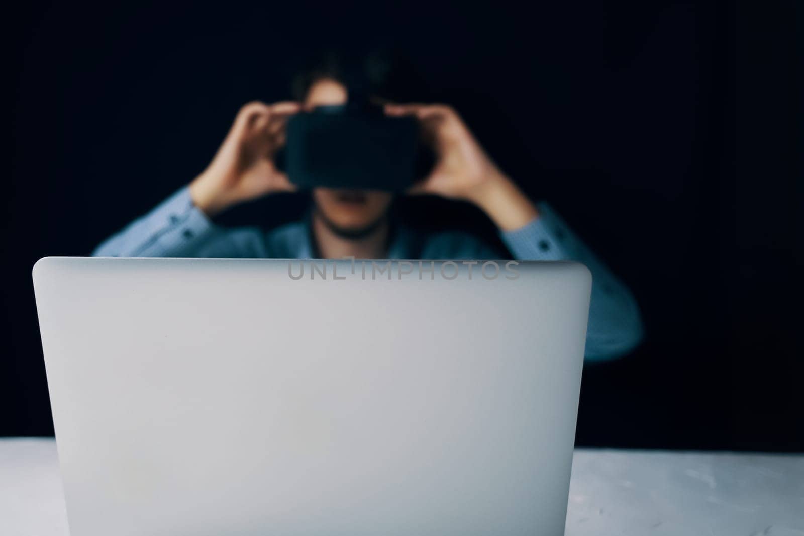 Blurred man in VR headset operating laptop in dark setting, depicting virtual reality technology use.