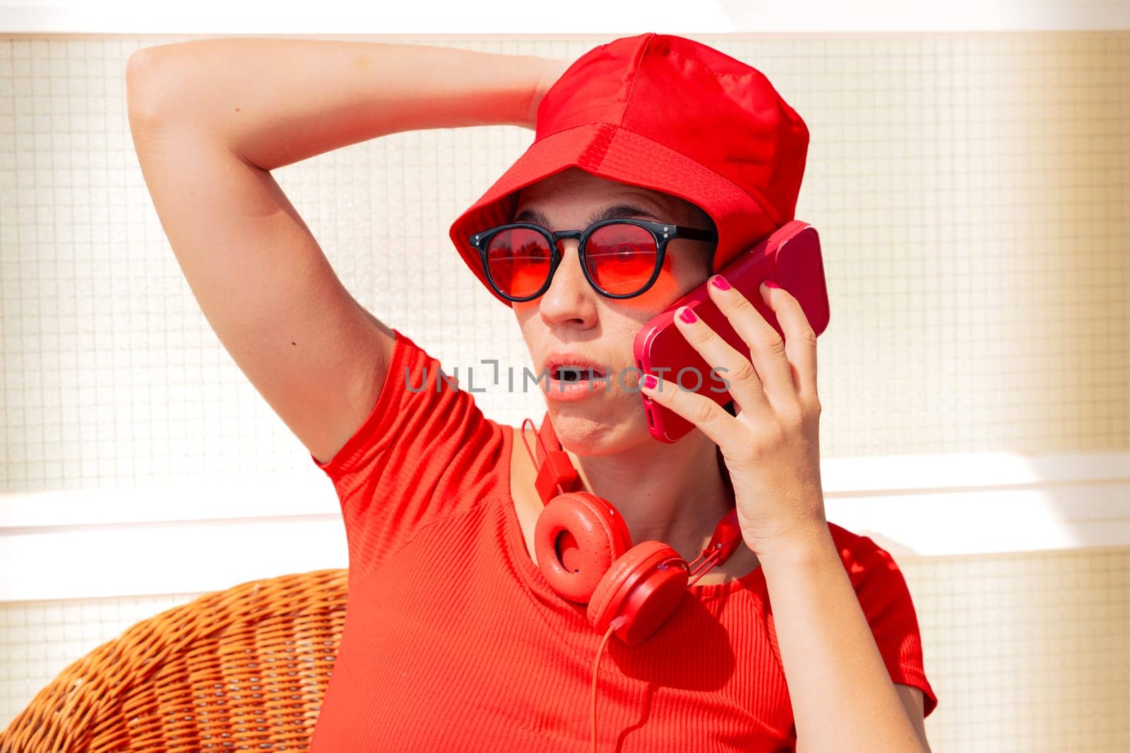 Young woman wearing sunglasses and red dress covering her mouth with her hand when talking on the phone with a friend and laughing