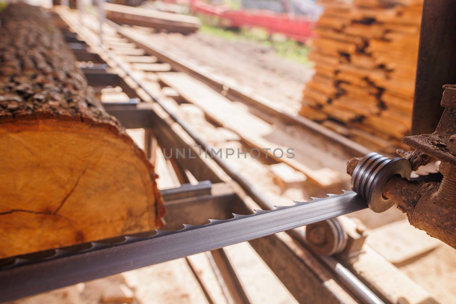 A close-up view of a sawmill with a large log being cut by a band saw. The saw blade is in focus, showing the cutting mechanism, while the log and sawmill environment are blurred in the background.