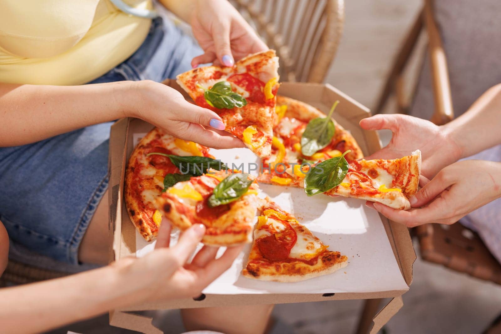 A group of friends share pizza outdoors, hands reaching for slices garnished with fresh basil, as a pizza box and plates of appetizers sit in the background.
