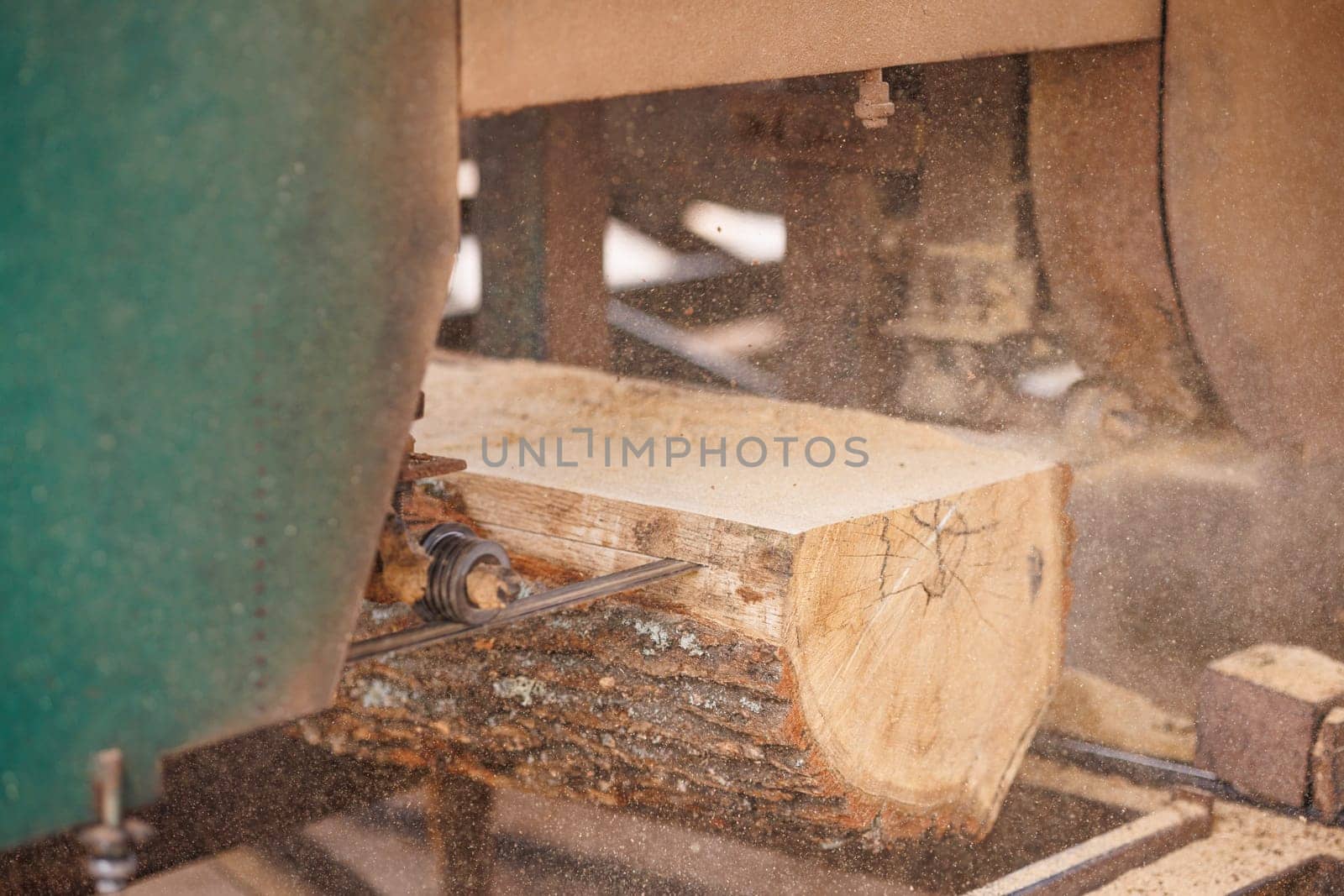 A close-up view of a sawmill with a large log being cut by a band saw. The saw blade is in focus, showing the cutting mechanism, while the log and sawmill environment are blurred in the background.