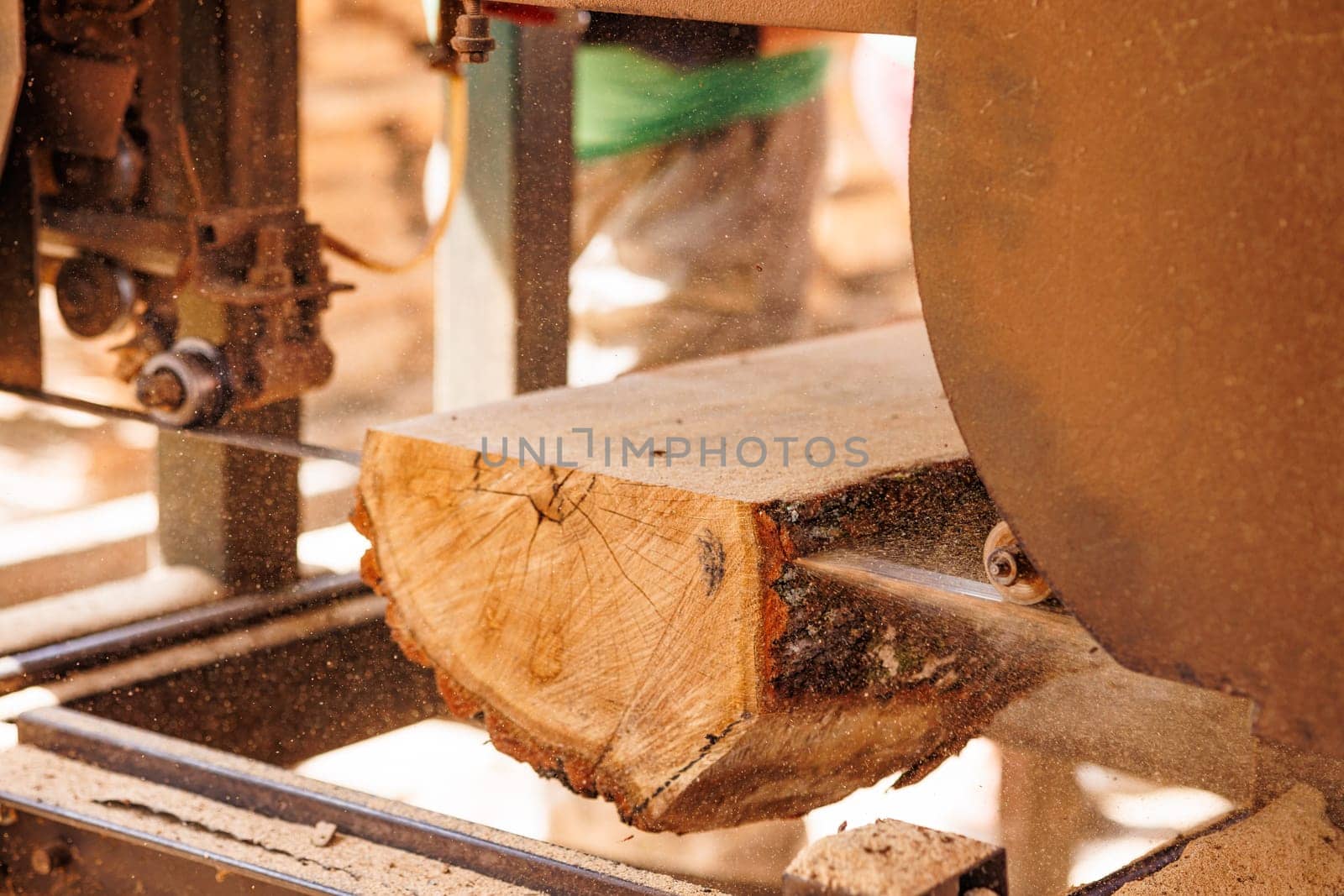 A close-up view of a log being cut on a sawmill. The saw blade is visible, and sawdust is flying around, indicating active cutting. The log shows natural wood grain and a rough texture.