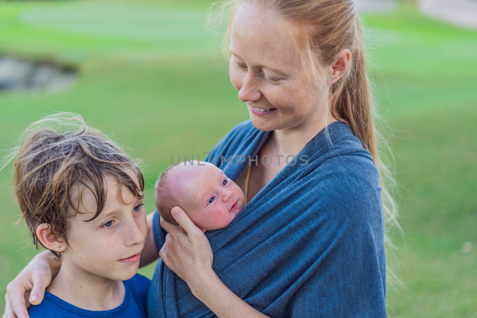 Mom walks with a newborn in a sling and also with her teenage son. This scene showcases the bond within the family, highlighting the nurturing care of a mother and the connection between siblings.