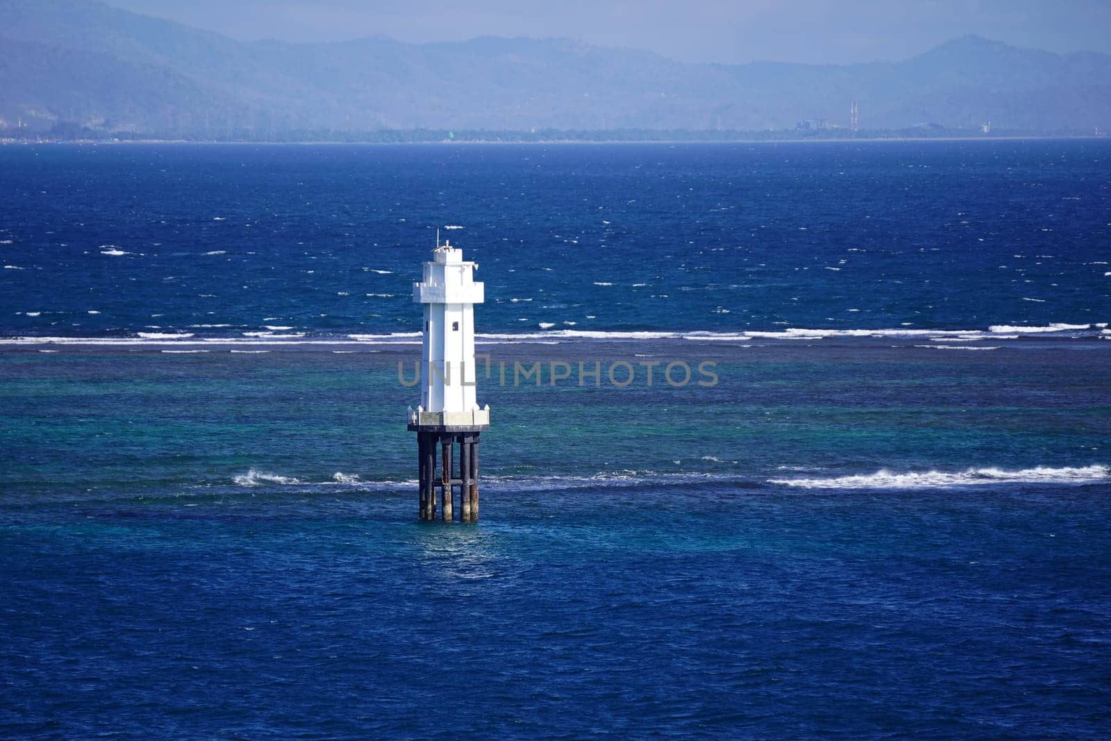 A solitary white lighthouse stands tall on a rocky outcrop in the vast blue ocean, surrounded by turquoise shallows, near Pantai Senggigi, Lombok, Indonesia.