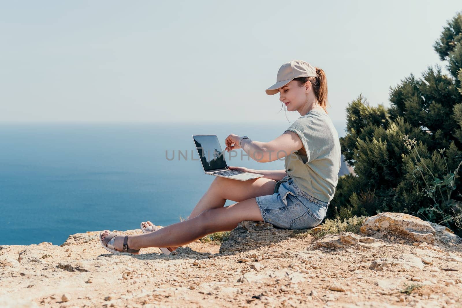 A young woman wearing a hat and casual clothes uses a laptop on a cliff with ocean views.