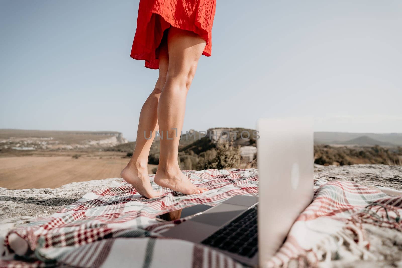 A woman in a red dress stands on a blanket with a laptop on it. The backdrop is a rocky cliff.