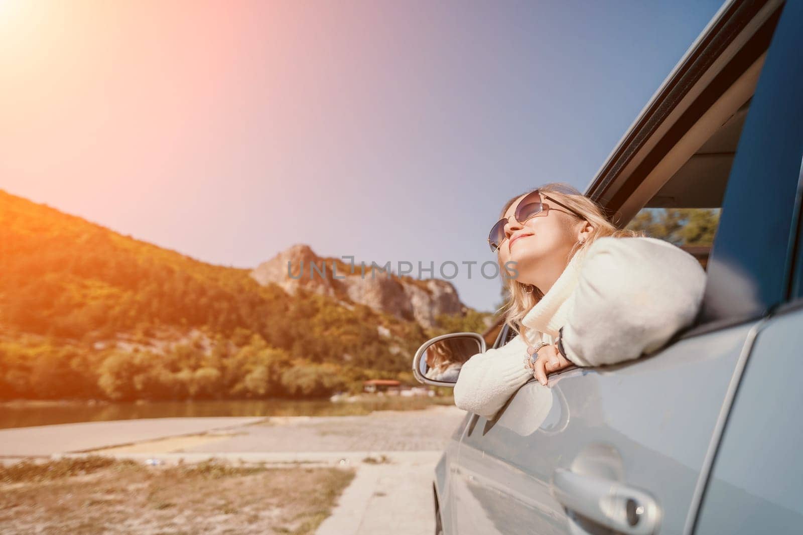 A woman in sunglasses leans out of a car window, enjoying the view of mountains and a winding road.