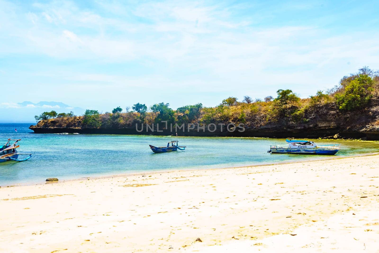 scene of a pristine beach near Pink Beach in Lombok, Indonesia. Clear turquoise waters lap against the white sandy shore, while colorful fishing boats bob gently in the bay