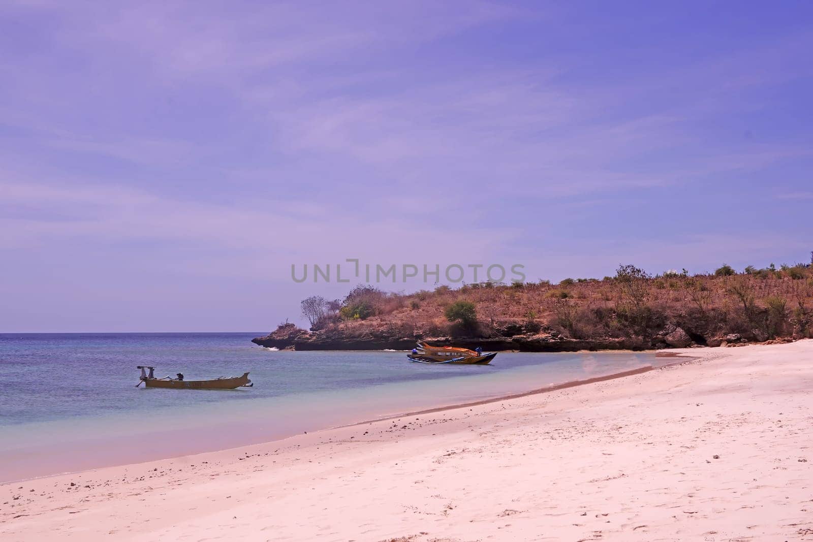 scene of a pristine beach near Pink Beach in Lombok, Indonesia. Clear turquoise waters lap against the white sandy shore, while colorful fishing boats bob gently in the bay