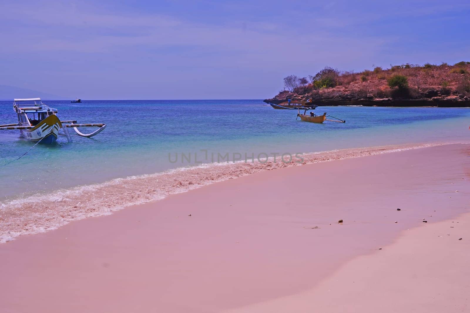 scene of a pristine beach near Pink Beach in Lombok, Indonesia. Clear turquoise waters lap against the white sandy shore, while colorful fishing boats bob gently in the bay
