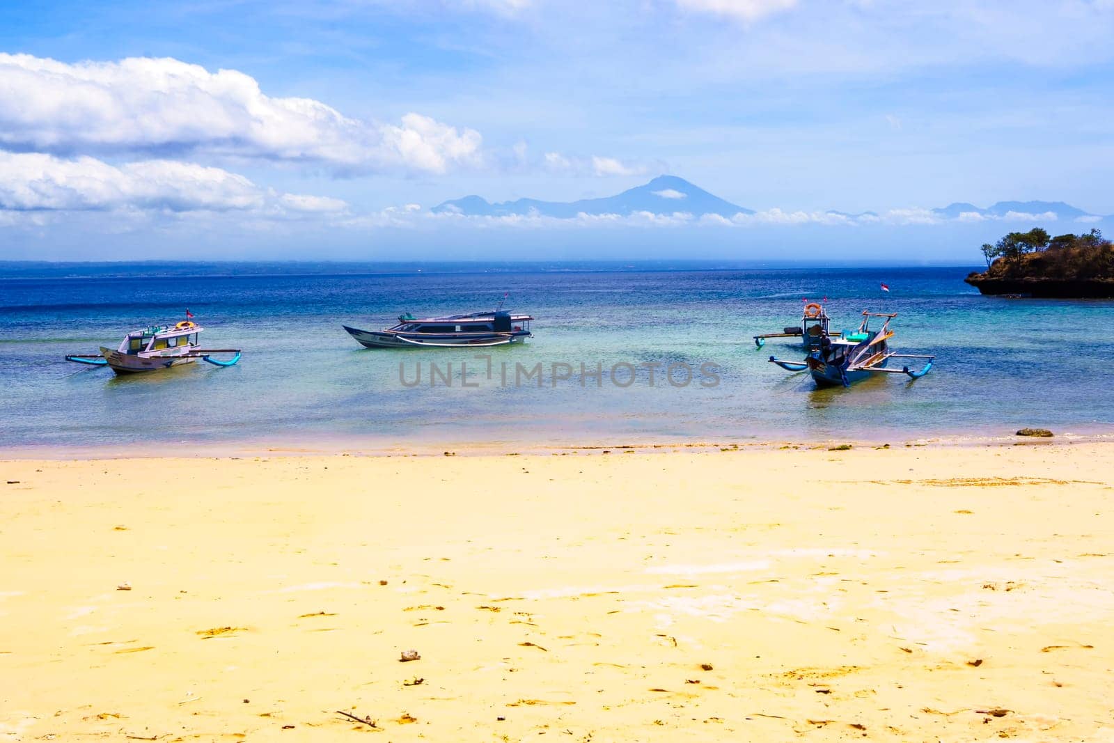 scene of a pristine beach near Pink Beach in Lombok, Indonesia. Clear turquoise waters lap against the white sandy shore, while colorful fishing boats bob gently in the bay