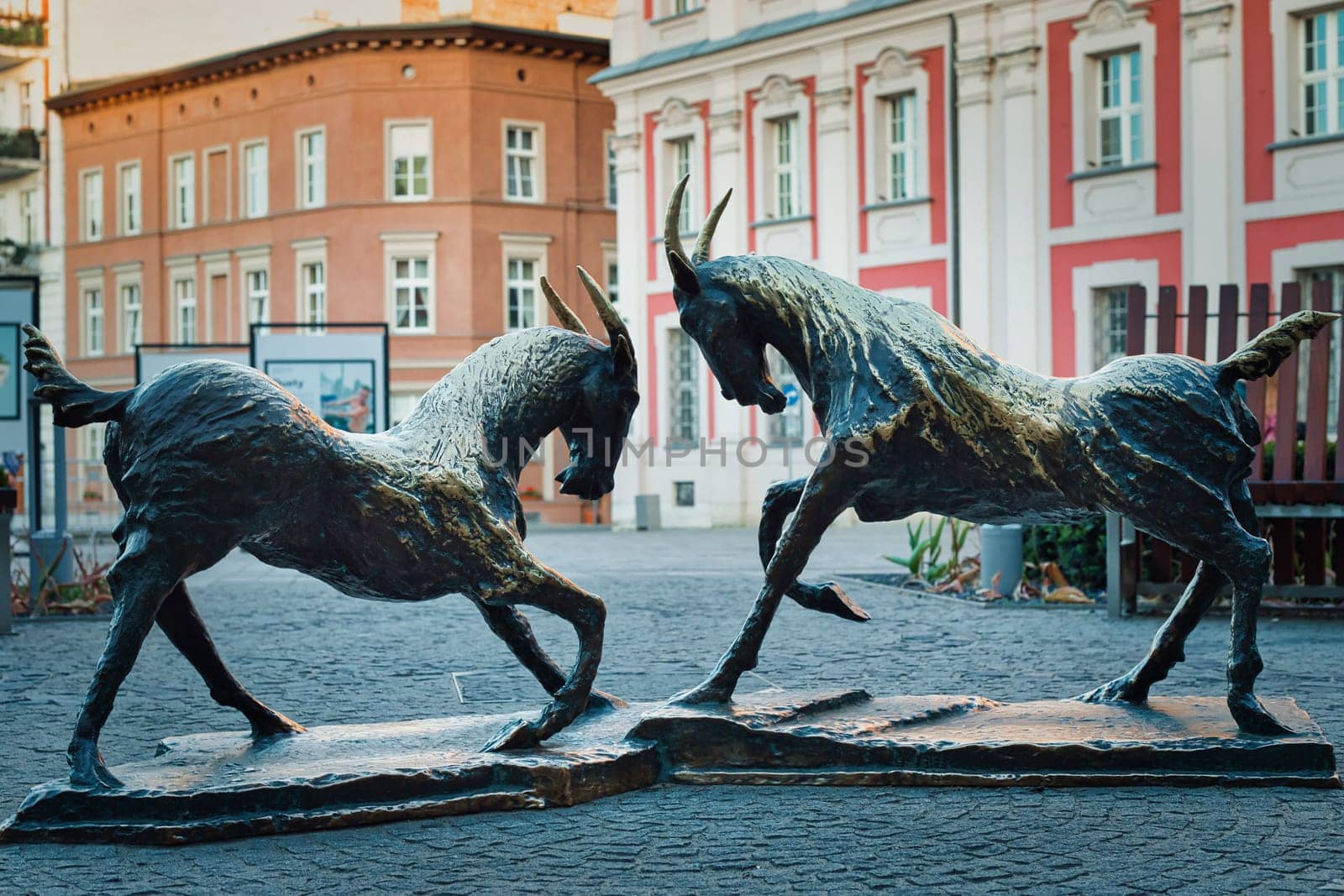 An artistic depiction of two bronze goat statues in a dynamic pose, located in the historic city square of Poznan, Poland. These sculptures symbolize the famous Poznan goats, a well known cultural emblem of the city. The statues are set against the backdrop of classic European architecture, adding to the charm of this heritage site. Ideal for themes related to travel, culture, history, and European landmarks