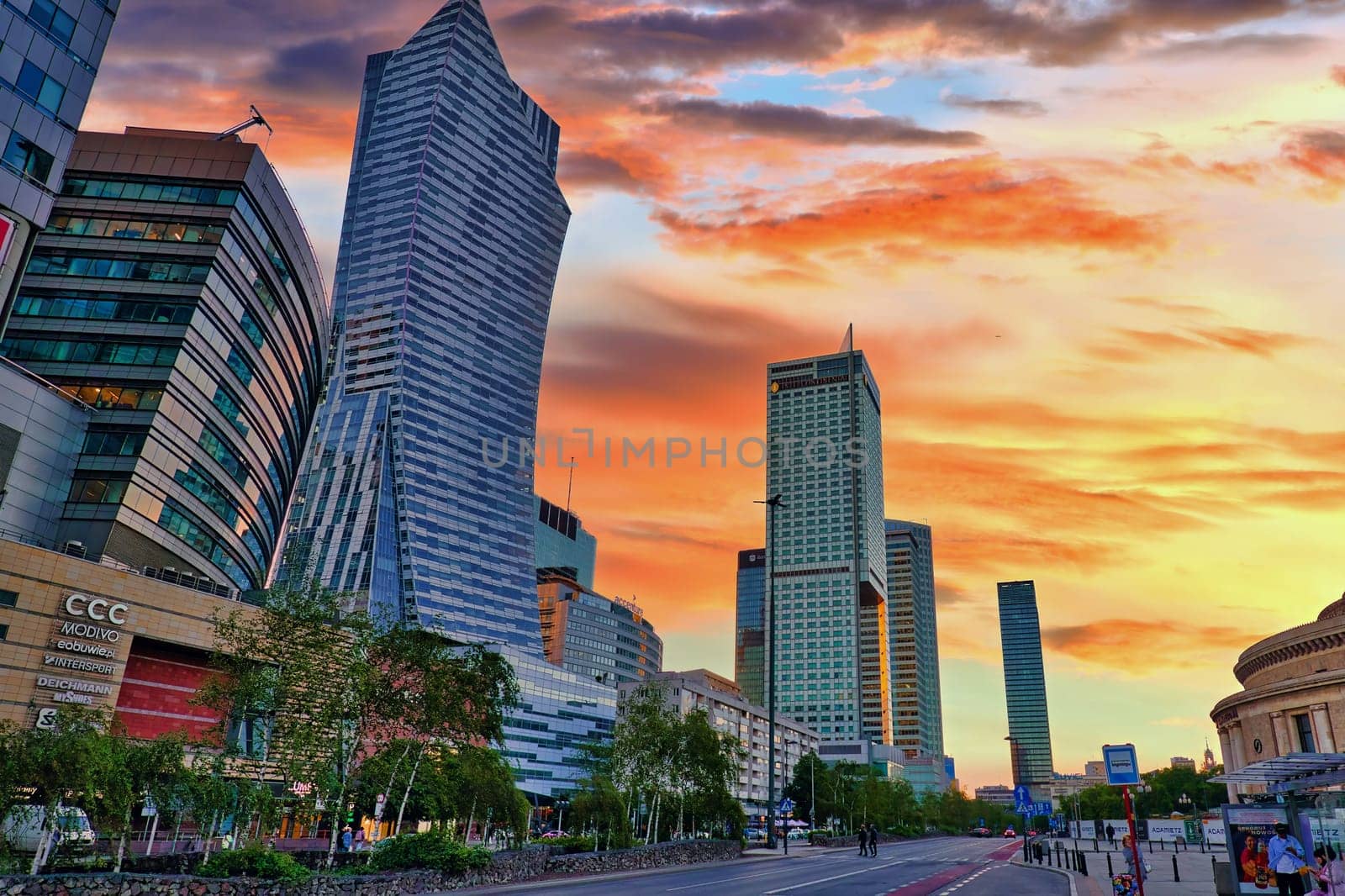 A stunning image of Warsaw's skyline featuring modern skyscrapers against a colorful sunset sky. The dynamic architecture, including curved and towering buildings, highlights the contemporary urban development of Poland capital. The warm hues of the sky contrast beautifully with the sleek designs of the buildings, creating an eye-catching cityscape. Ideal for use in travel, architecture, and cityscape themes.