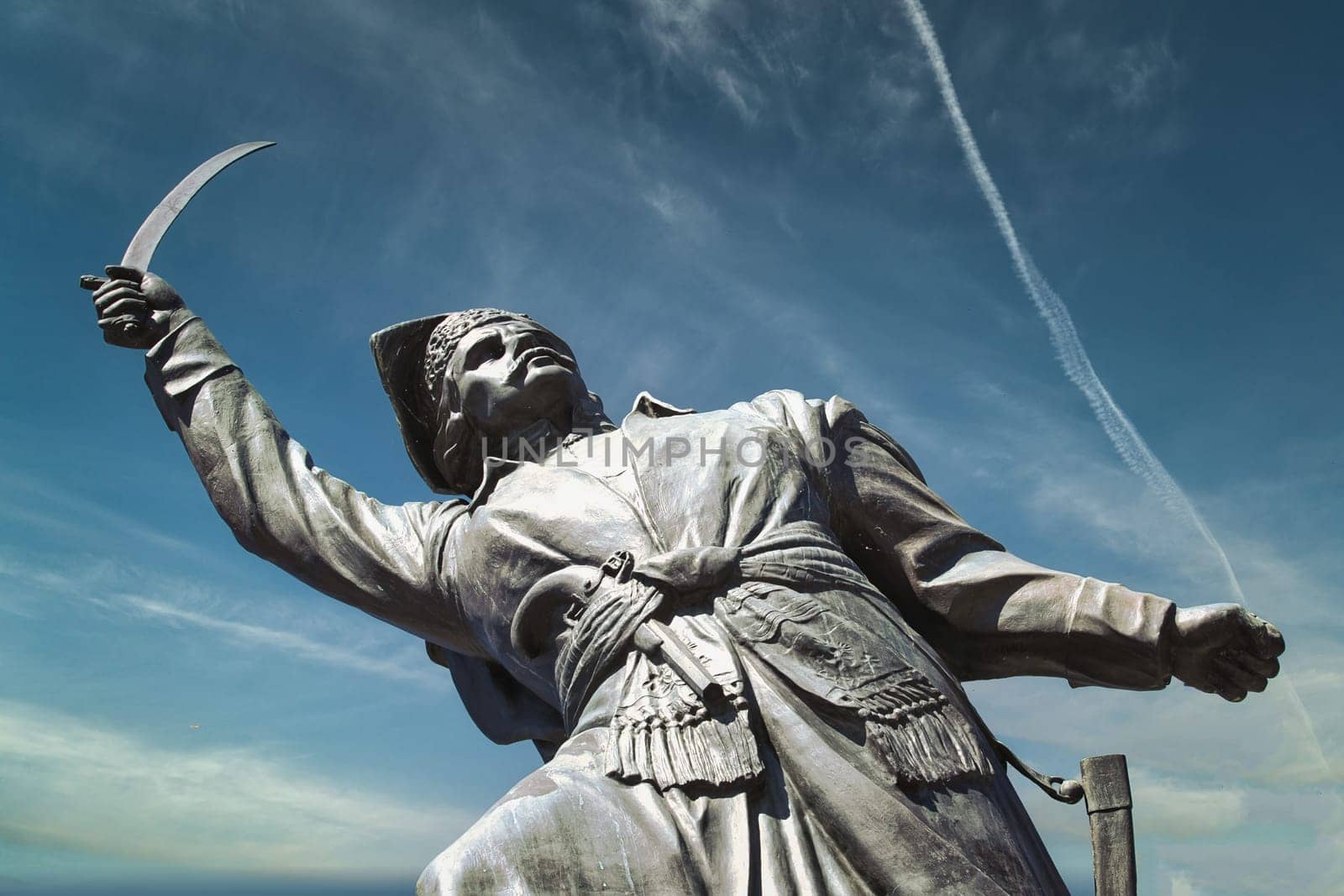 A striking image of a historic statue of a warrior brandishing a sword against a vivid blue sky. The powerful stance and detailed craftsmanship of the statue convey strength and determination, making it a prominent cultural landmark. Captured with a low-angle perspective, the statue symbolizes bravery and heroism. Perfect for use in historical, cultural, and travel-related themes.