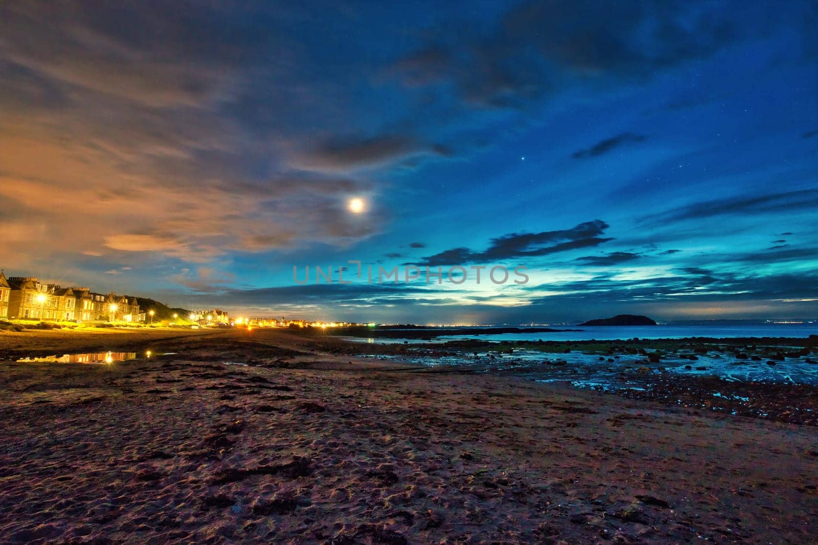 A breathtaking view of North Berwick Beach in the United Kingdom captured during dusk. The image showcases a vibrant sky transitioning into night with shades of blue, orange, and purple, while city lights illuminate the shoreline in the distance. The calm sea reflects the evening sky, creating a serene and captivating scene. Perfect for themes related to coastal landscapes, twilight photography, and travel.